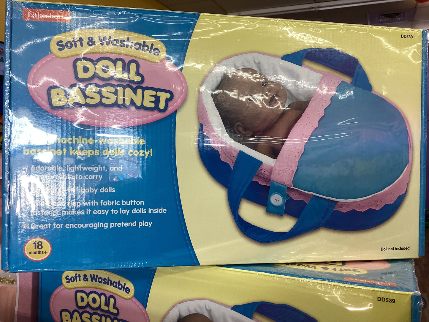Packaged doll bassinet in pink and blue with a fabric handle, displayed in a retail setting.